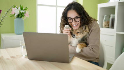 Young hispanic woman with dog sitting on table having video call at dinning room