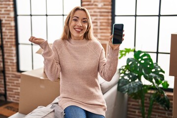 Hispanic woman moving to a new home showing smartphone screen screaming proud, celebrating victory and success very excited with raised arm