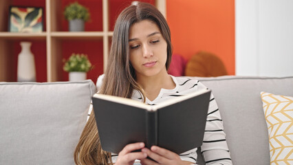 Young beautiful hispanic woman reading book sitting on sofa at home