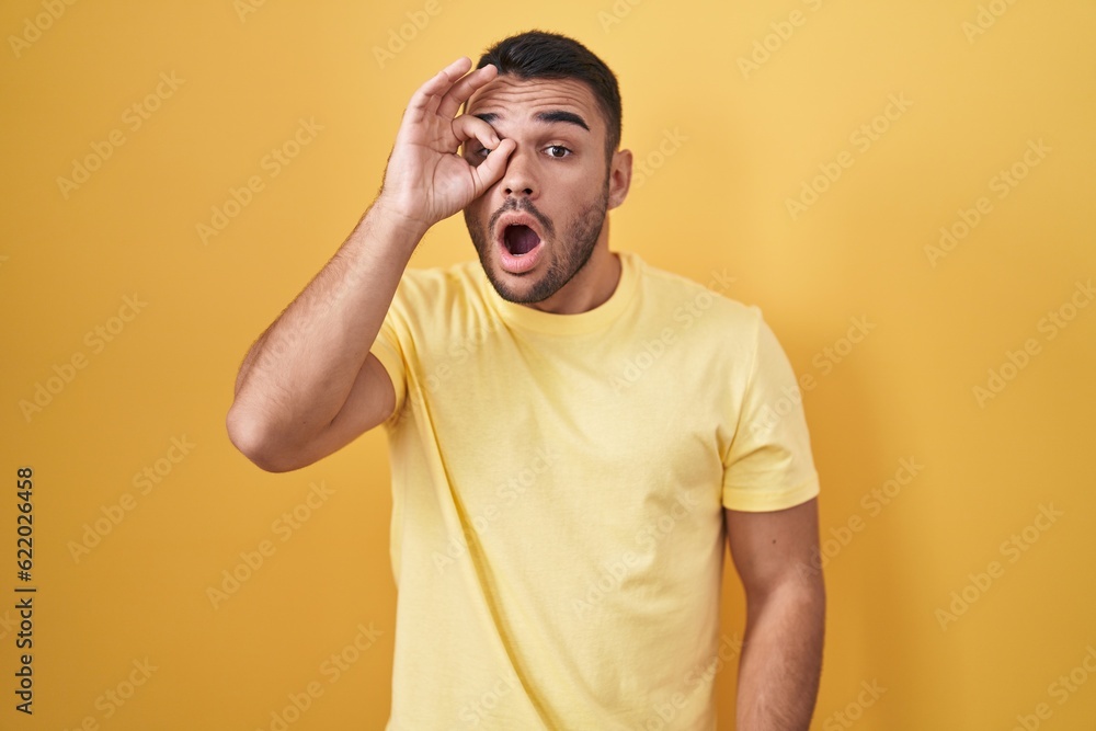 Sticker young hispanic man standing over yellow background doing ok gesture shocked with surprised face, eye