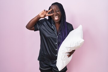 Young african woman wearing pijama hugging pillow doing peace symbol with fingers over face,...