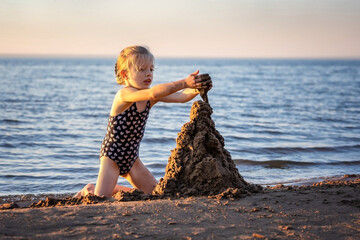 Little girl builds a sand castle on the beach. A child plays in the sand on the river bank. Summer children's games on the sea.