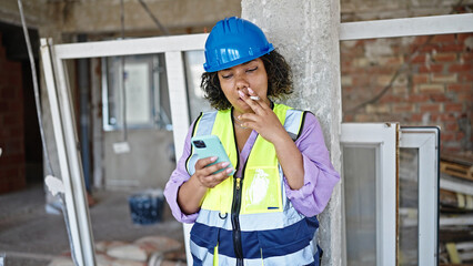 Young beautiful latin woman builder smoking cigarette using smartphone at construction site