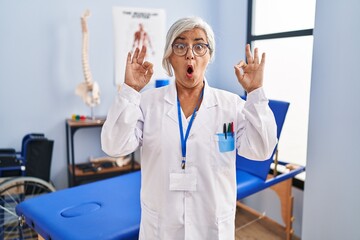 Middle age woman with grey hair working at pain recovery clinic looking surprised and shocked doing ok approval symbol with fingers. crazy expression