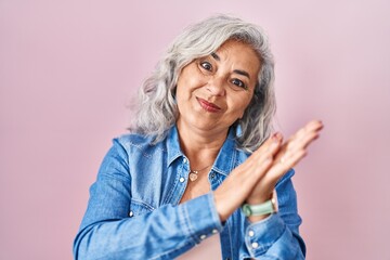 Middle age woman with grey hair standing over pink background clapping and applauding happy and joyful, smiling proud hands together