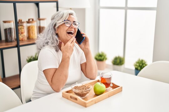 Middle age woman having breakfast talking on the smartphone at home