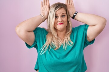 Caucasian plus size woman standing over pink background doing bunny ears gesture with hands palms looking cynical and skeptical. easter rabbit concept.