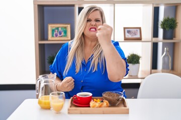 Obraz na płótnie Canvas Caucasian plus size woman eating breakfast at home angry and mad raising fist frustrated and furious while shouting with anger. rage and aggressive concept.