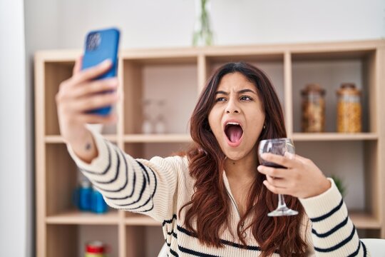 Hispanic young business woman taking a selfie picture drinking a glass of wine angry and mad screaming frustrated and furious, shouting with anger. rage and aggressive concept.