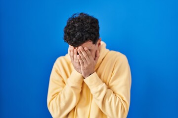 Hispanic man standing over blue background with sad expression covering face with hands while crying. depression concept.