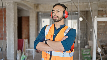 Young hispanic man builder with crossed arms wearing noise cancelling earmuffs at construction site