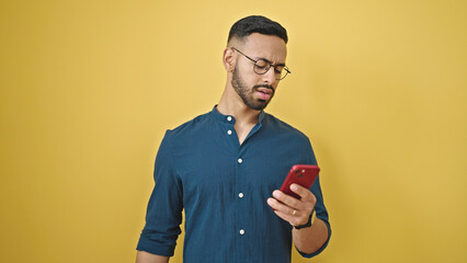 Young hispanic man using smartphone upset over isolated yellow background