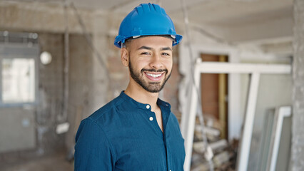 Young hispanic man architect wearing hardhat smiling at construction site