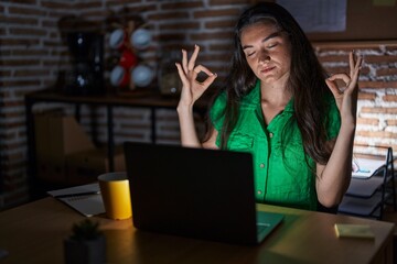 Young teenager girl working at the office at night relax and smiling with eyes closed doing meditation gesture with fingers. yoga concept.