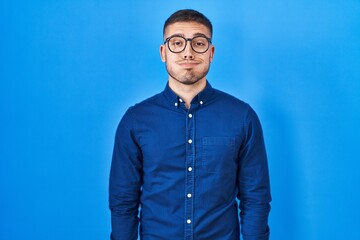 Young hispanic man wearing glasses over blue background puffing cheeks with funny face. mouth...
