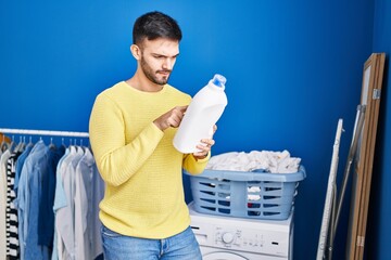 Young hispanic man reading detergent label with relaxed expression at laundry room
