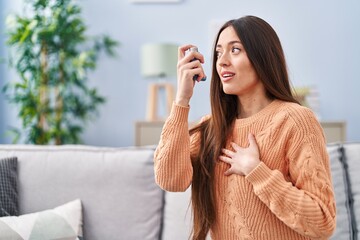 Young beautiful hispanic woman using inhaler sitting on sofa at home