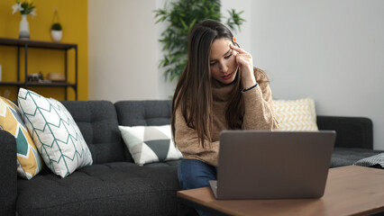 Beautiful hispanic woman using laptop sitting on sofa with worried expression at home