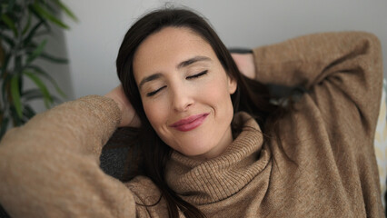 Beautiful hispanic woman relaxed with hands on head sitting on sofa at home