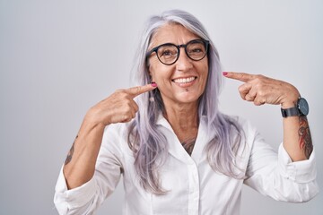 Middle age woman with tattoos wearing glasses standing over white background smiling cheerful showing and pointing with fingers teeth and mouth. dental health concept.