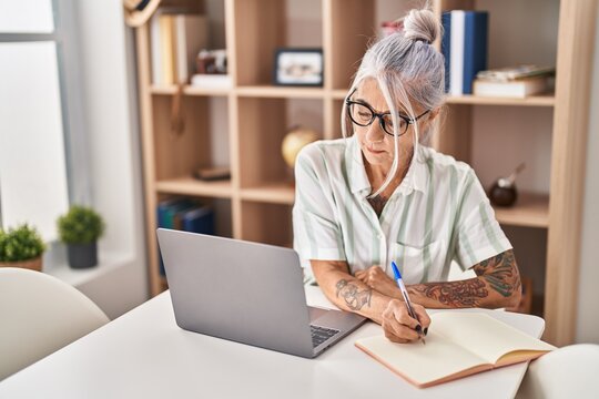 Middle age grey-haired woman writing on notebook using laptop at home