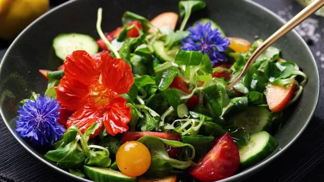 Eating with fork green salad with fresh cucumbers, tomato, peach and nasturtium flowers on black background. Close up