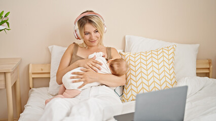 Mother and daughter sitting on bed breastfeeding baby listening to music at bedroom