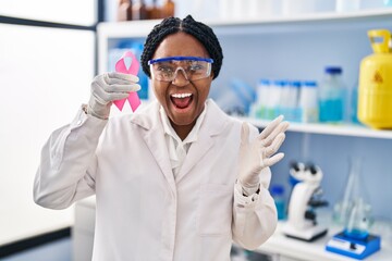 African american woman working at scientist laboratory holding pink ribbon celebrating victory with...