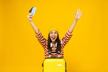 Happy young woman in striped t-shirt with suitcase and tickets on yellow background