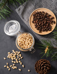 Pine nuts in a jar, bowl and a scattered on a dark background with branches of pine needles and cone. The concept of a natural, organic and healthy superfood and snack.