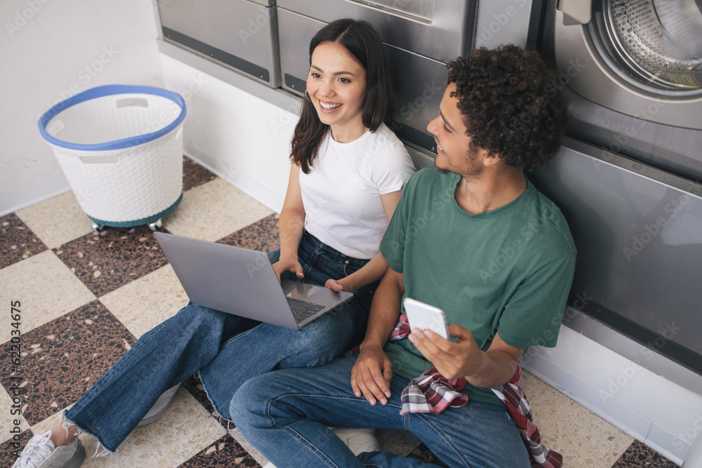 Wall mural cheerful students couple with laptop and smartphone sitting at laundrette