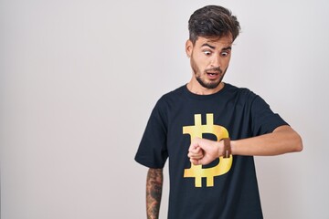 Young hispanic man with tattoos wearing bitcoin t shirt looking at the watch time worried, afraid...