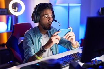 Young hispanic man with tattoos playing video games with smartphone scared and amazed with open mouth for surprise, disbelief face - obrazy, fototapety, plakaty
