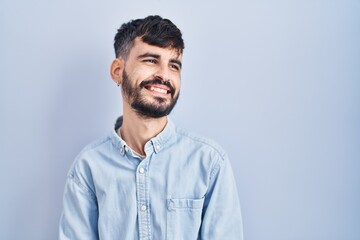 Fototapeta premium Young hispanic man with beard standing over blue background looking away to side with smile on face, natural expression. laughing confident.