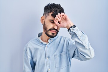 Young hispanic man with beard standing over blue background touching forehead for illness and fever, flu and cold, virus sick