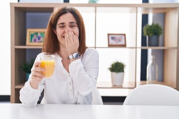 Brunette woman drinking glass of orange juice laughing and embarrassed giggle covering mouth with hands, gossip and scandal concept