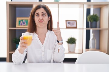 Brunette woman drinking glass of orange juice pointing up looking sad and upset, indicating direction with fingers, unhappy and depressed.