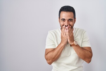 Hispanic man with beard standing over isolated background laughing and embarrassed giggle covering...