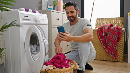 Young hispanic man using smartphone washing clothes smiling at laundry room