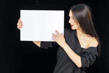 attractive girl in dark clothes holds an empty billboard in her hands on a dark background and points to it, attracting attention