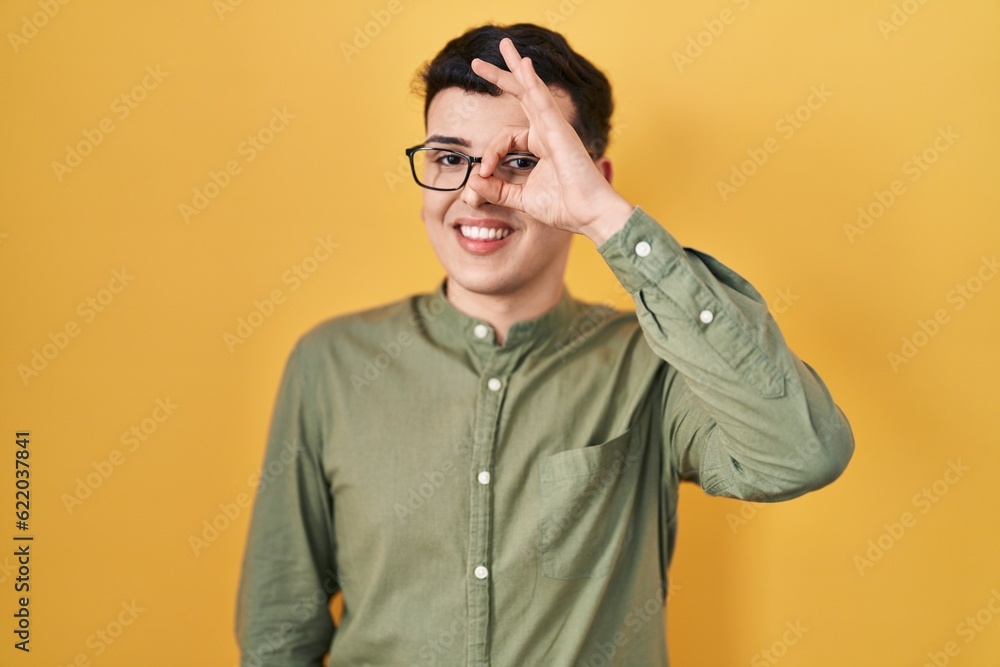 Wall mural non binary person standing over yellow background doing ok gesture with hand smiling, eye looking th