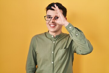 Fototapeta premium Non binary person standing over yellow background doing ok gesture with hand smiling, eye looking through fingers with happy face.