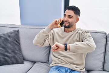 Young arab man talking on the smartphone looking watch at home