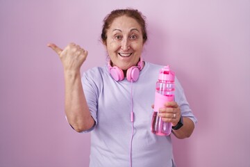 Senior woman wearing sportswear and headphones pointing to the back behind with hand and thumbs up, smiling confident