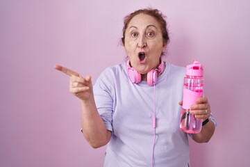 Senior woman wearing sportswear and headphones surprised pointing with finger to the side, open mouth amazed expression.