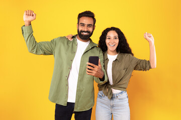 Arab couple celebrating holding smarphone and shaking fists, yellow background