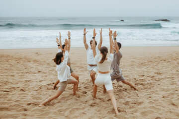 Group of young men and women in sportswear doing yoga on ocean shore, standing on mats in circle,...