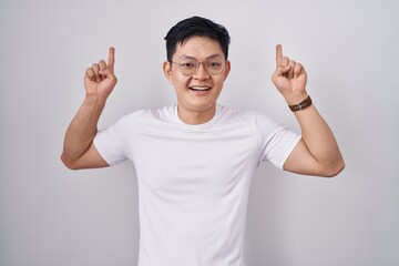 Young asian man standing over white background smiling amazed and surprised and pointing up with fingers and raised arms.