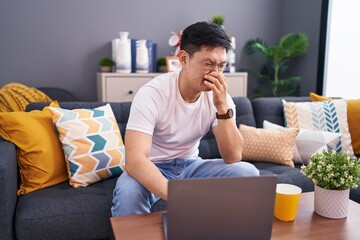 Young asian man using laptop at home sitting on the sofa smelling something stinky and disgusting, intolerable smell, holding breath with fingers on nose. bad smell - obrazy, fototapety, plakaty