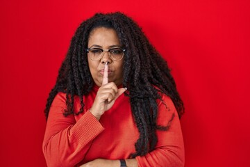 Plus size hispanic woman standing over red background asking to be quiet with finger on lips. silence and secret concept.